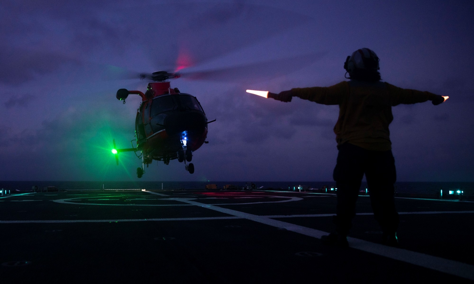 Landing Signals Officer Ensign directs an MH-65E Dolphin helicopter during routine nighttime flight operations onboard U.S. Coast Guard Cutter Munro (WMSL 755) Aug. 23, 2023, in the South China Sea. Munro deployed to the Western Pacific to conduct engagements with regional allies and partner nations, reinforcing a rules-based order in the maritime domain.  (U.S. Navy photo by Chief Petty Officer Brett Cote.)